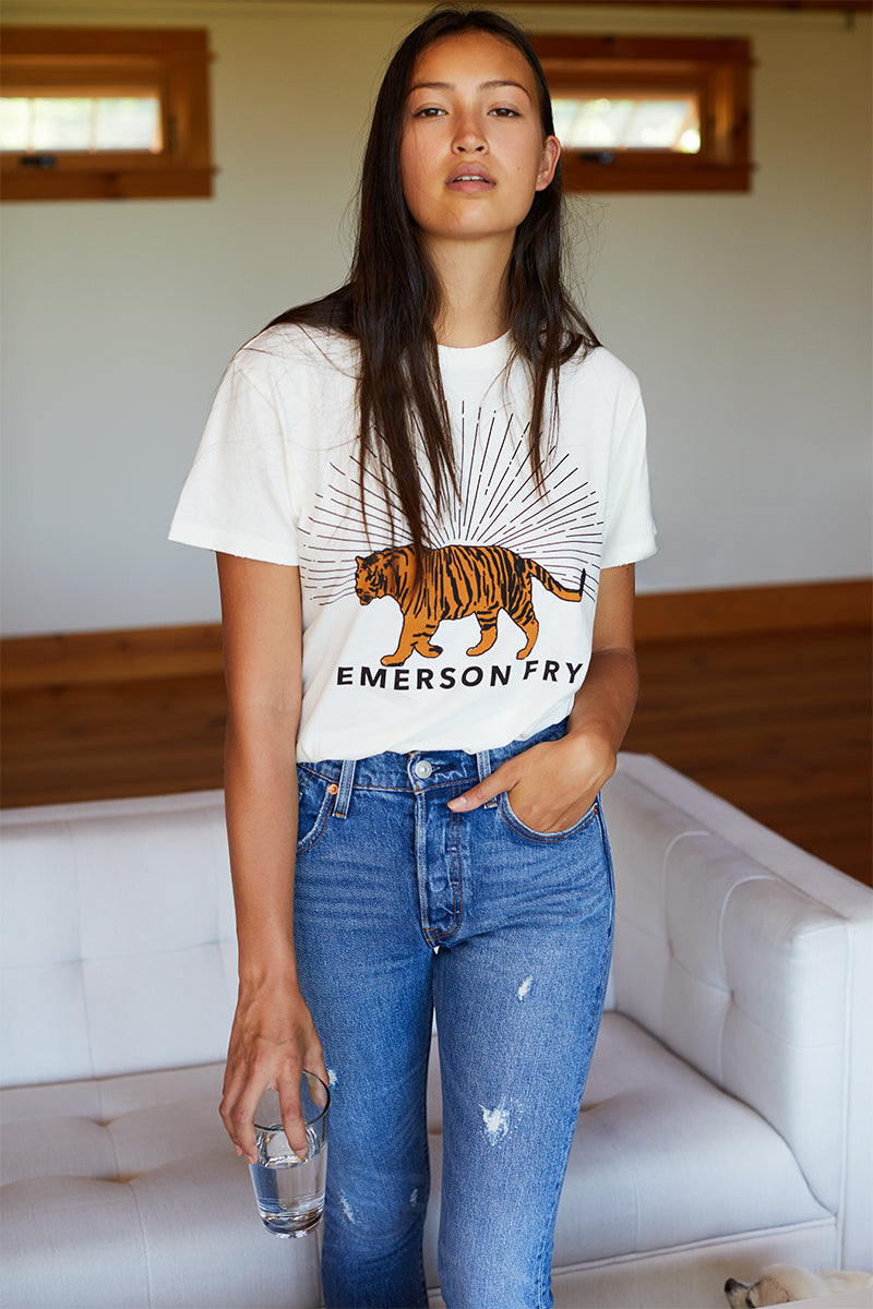 A woman wearing a white crew neck t-shirt with a graphic print of a tiger and the brand name 'EMERSON FRY' on the front. They are also wearing blue denim jeans.